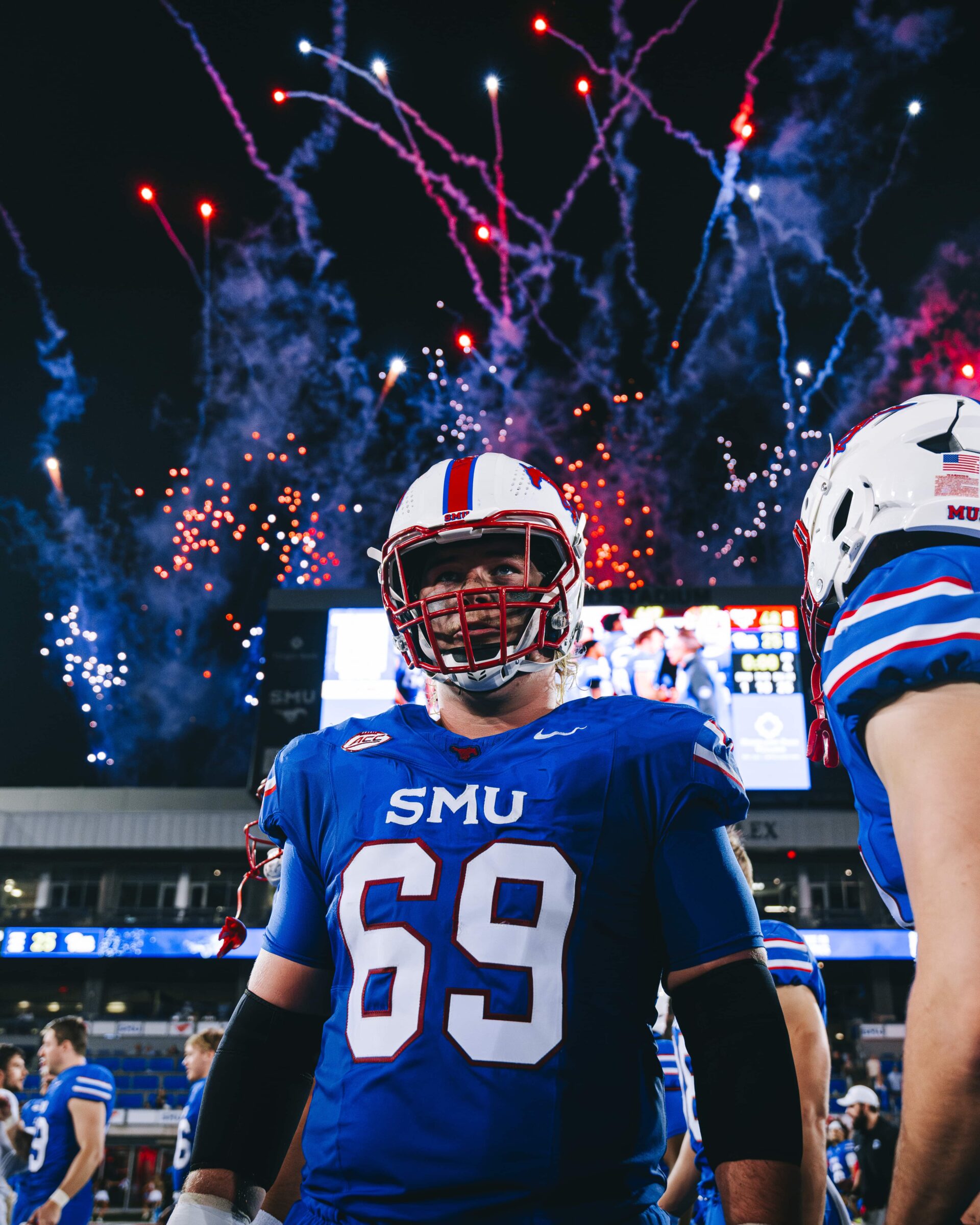 An SMU football player stands on the field as blue and red fireworks launch into the night sky behind him.