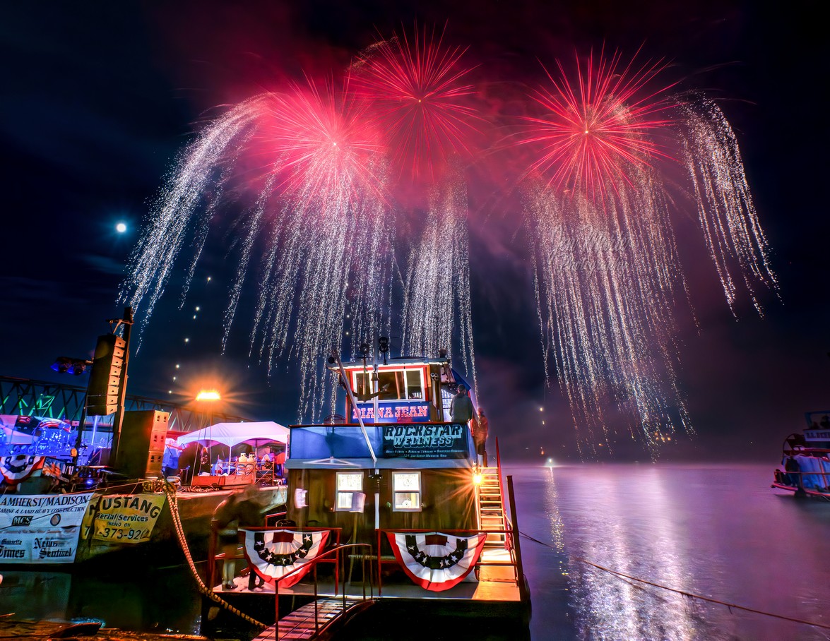 Red aerial fireworks cascade above illuminated riverboats on the Ohio River during the Sternwheel Festival, reflecting across the water.