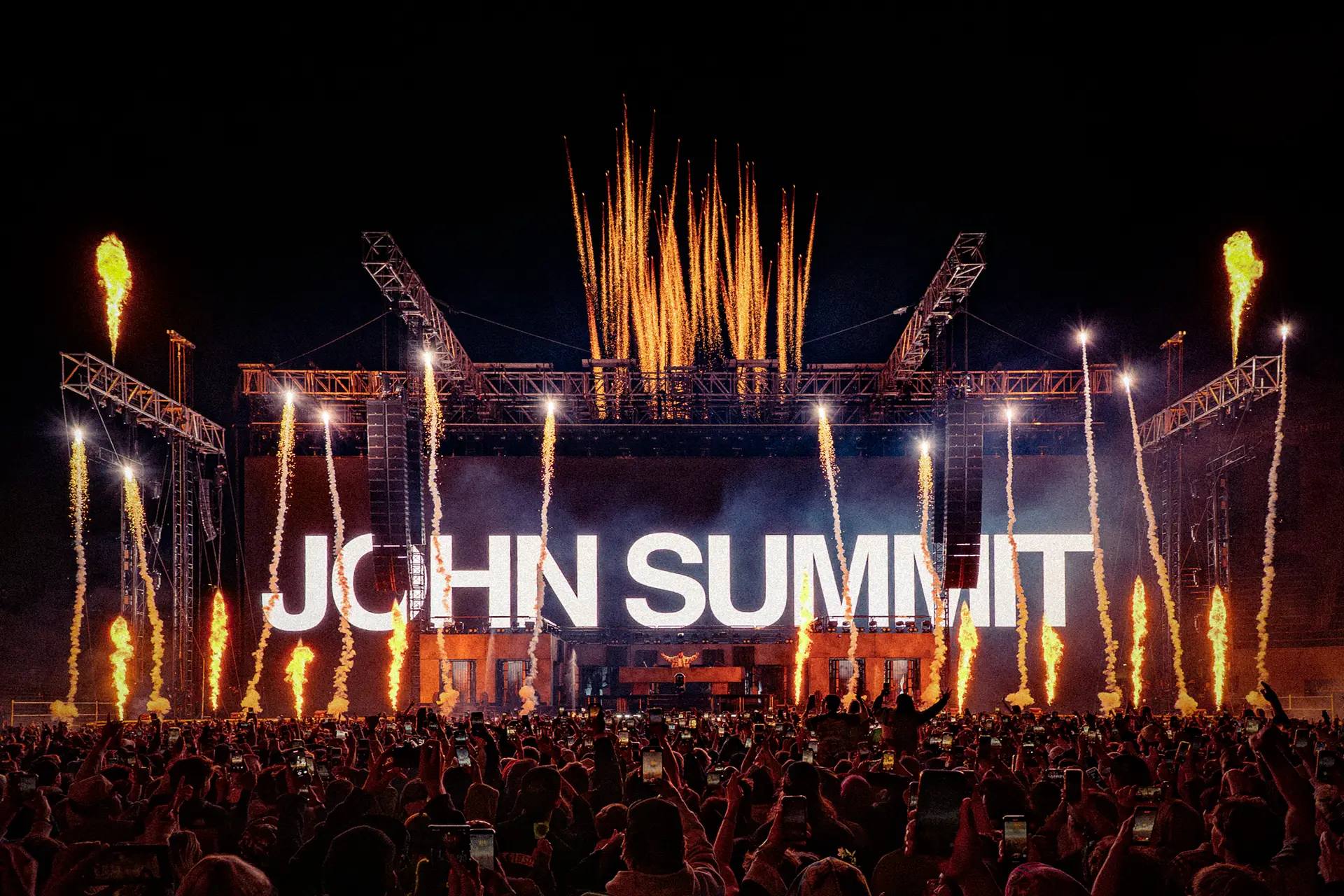 John Summit stands onstage at Folsom Field with vertical flame jets and lighting effects illuminating the performance behind him.