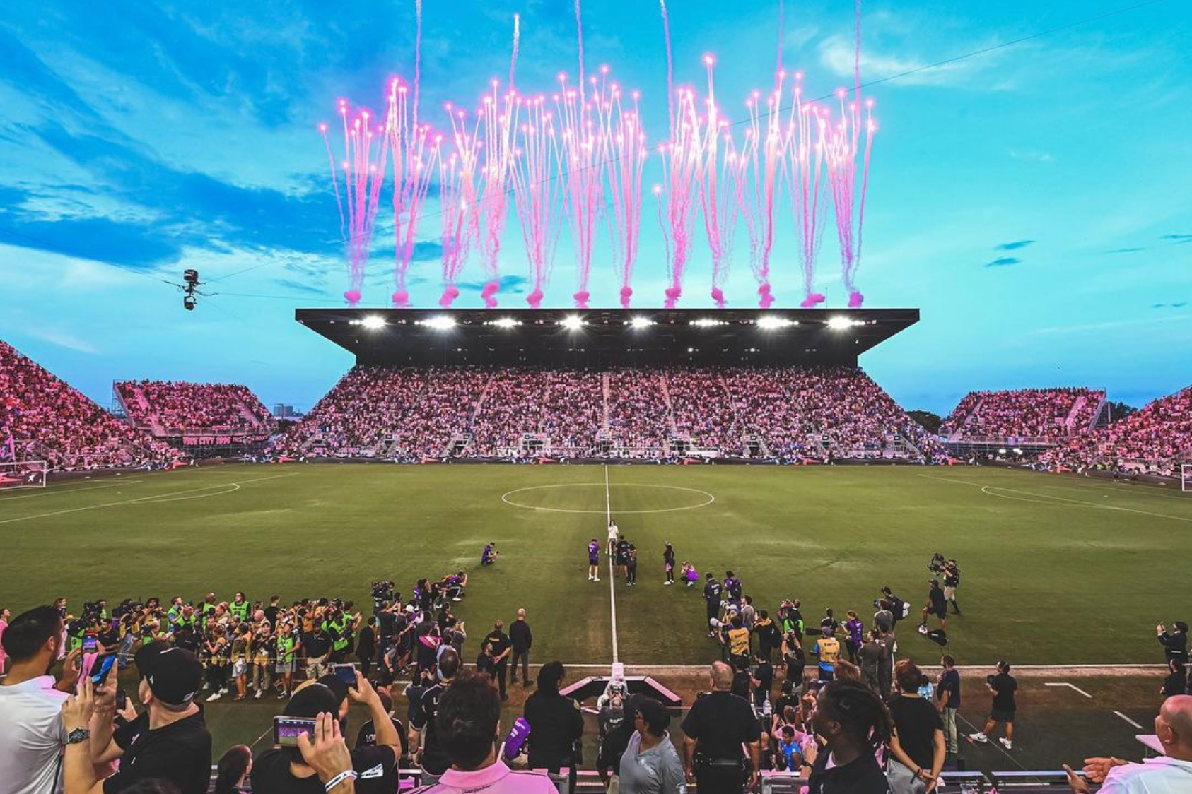 Pink aerial fireworks launch above the stadium roof as Inter Miami CF players line up on the field before a packed crowd.