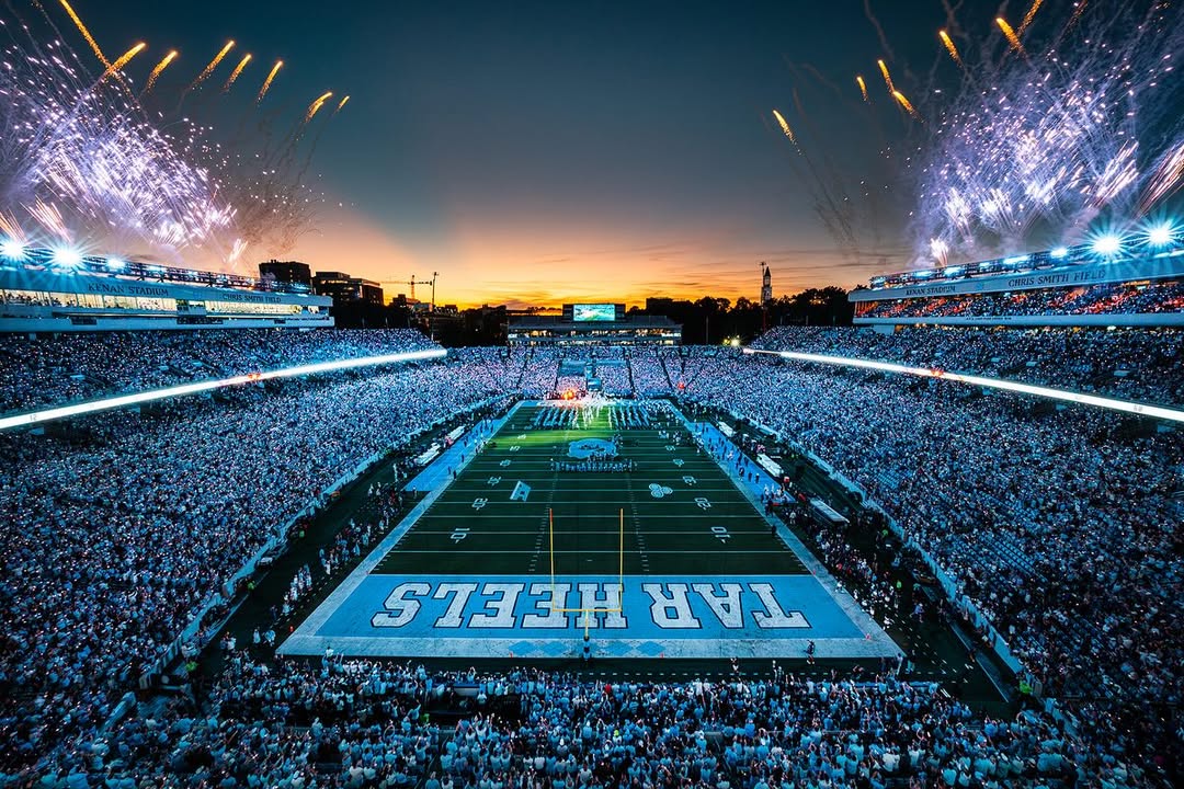 A packed UNC football stadium glows at dusk as aerial fireworks burst above the field during a Tar Heels home game introduction.