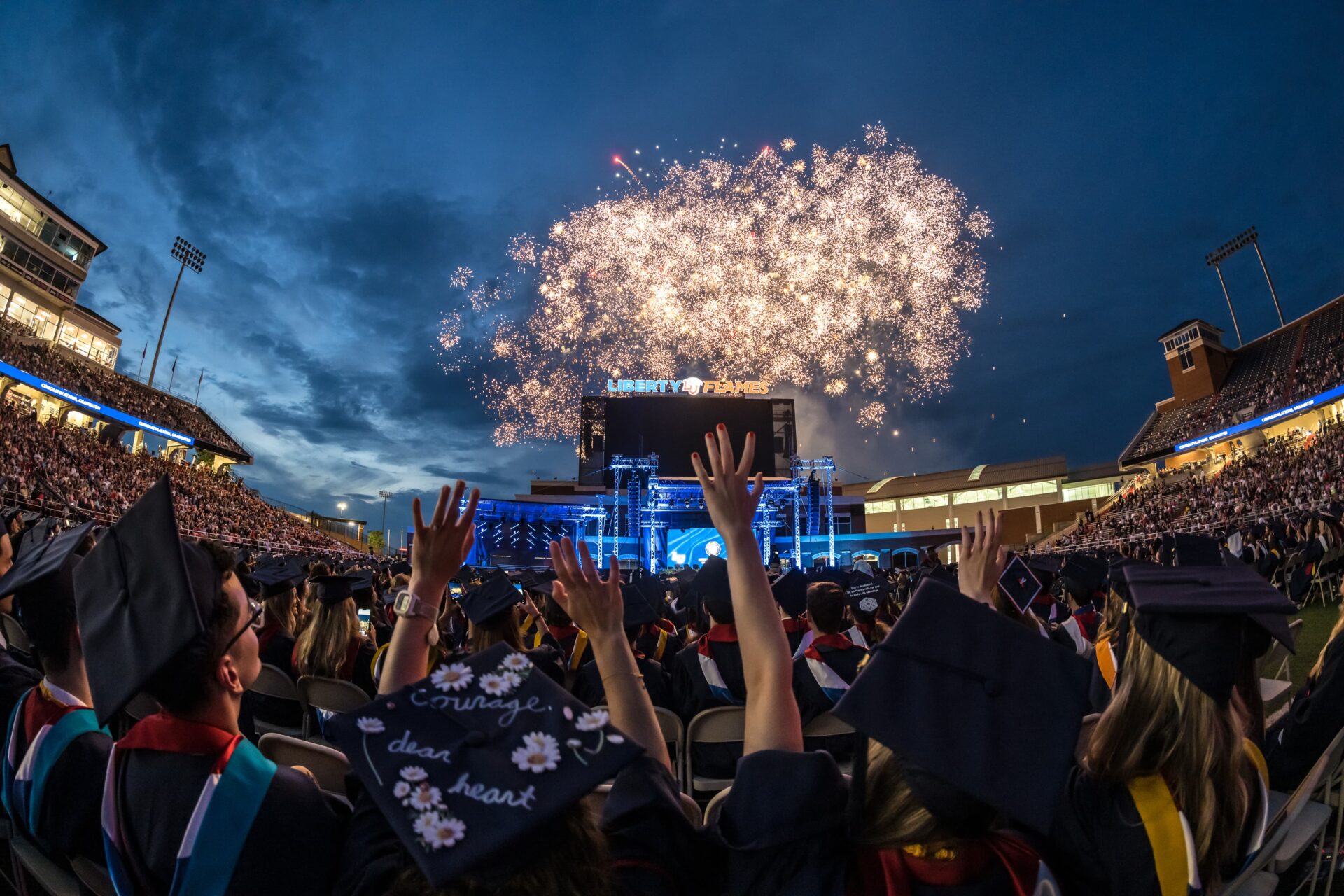 Large gold aerial fireworks and vertical spark effects illuminate graduates and guests at Liberty University’s commencement ceremony.